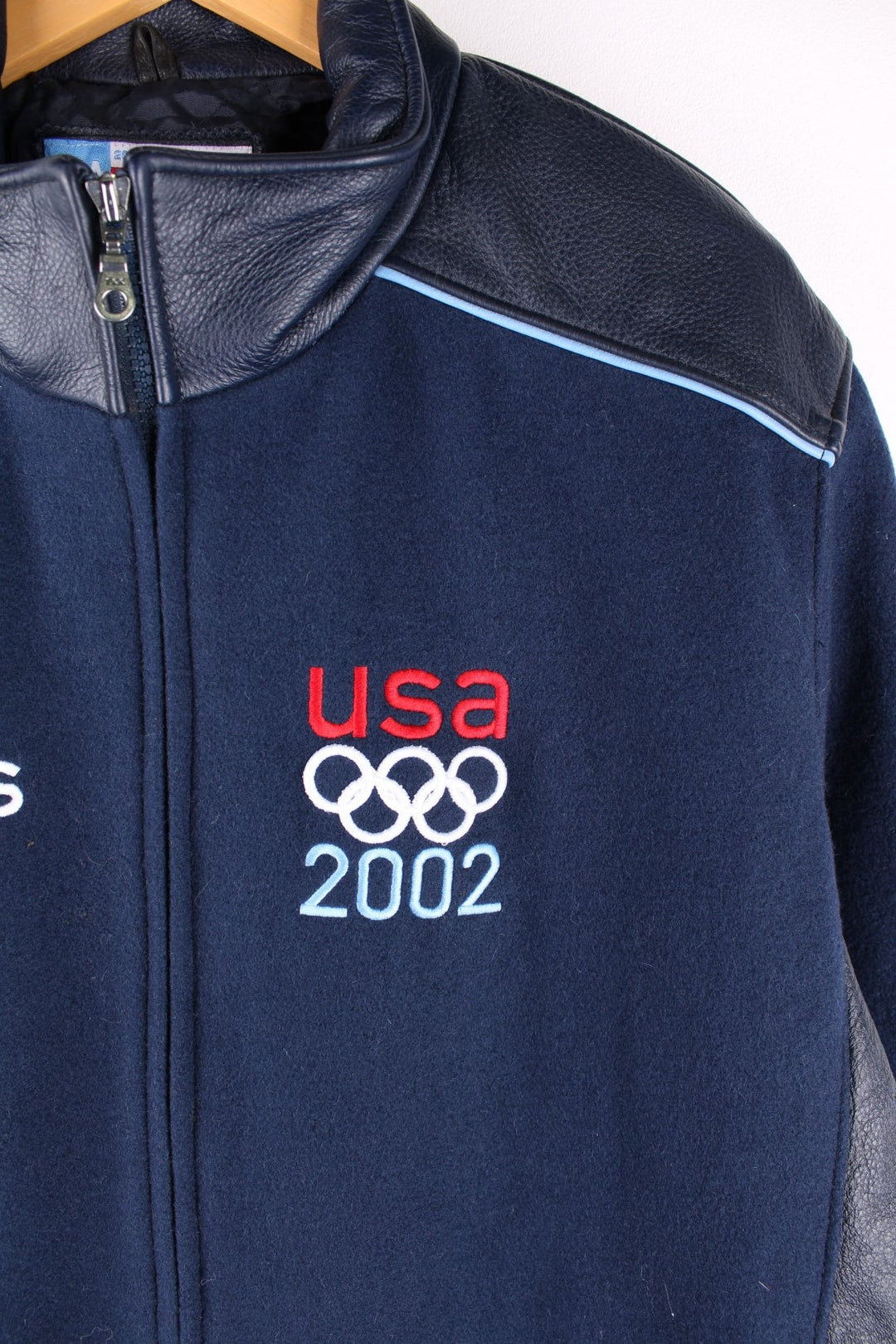 Vintage 2002 USA Winter Olympic Team Coat in a navy blue colourway. The jacket is wool, and has leather patches on the shoulders and side panels. There is a light blue piping on the shoulders too. There are multiple zip pockets on the front and sleeves, embroidered logo on the front, and a large USA flag embroidered on the back. The games took place in Salt Lake City Utah, and was the US's most successful winter games on record, winning 34 medals in total. 