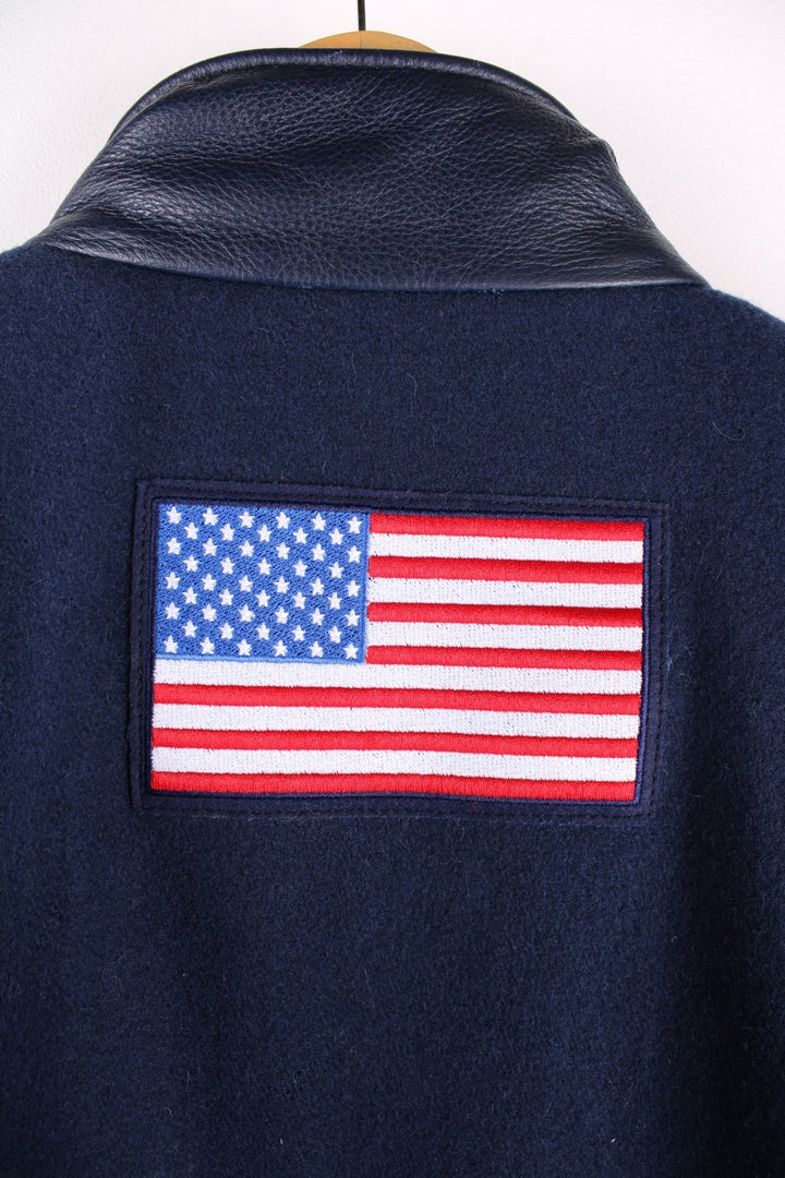 Vintage 2002 USA Winter Olympic Team Coat in a navy blue colourway. The jacket is wool, and has leather patches on the shoulders and side panels. There is a light blue piping on the shoulders too. There are multiple zip pockets on the front and sleeves, embroidered logo on the front, and a large USA flag embroidered on the back. The games took place in Salt Lake City Utah, and was the US's most successful winter games on record, winning 34 medals in total. 