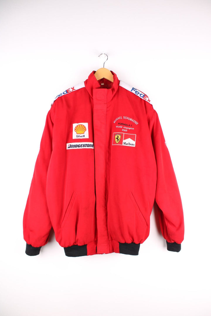 Vintage 2002 Michael Schumacher F1 souvenir jacket in a Ferrari red colourway. The jacket has multiple brand patches down the sleeves, Fed Ex shoulder lapels and a zip/velcro fastening to close. It has a large Marlboro/ Ferrari patch on the back which has to be the most iconic brand collaboration in F1 history. The jacket is dated to 2002, when Schumacher won 11 out of 17 races during the racing season and finished on the podium for all 17. 