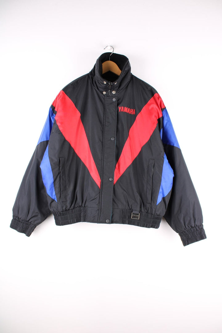 Vintage Yamaha motorcycle jacket in a black, blue and red colourway, the jacket is quilted, has an elasticated hem and interior pocket. It has flashes of red and blue and a large YAMAHA logo embroidered on the back. There is also a small metal Yamaha lgo pinned to the front. Dates to the 1990s. 