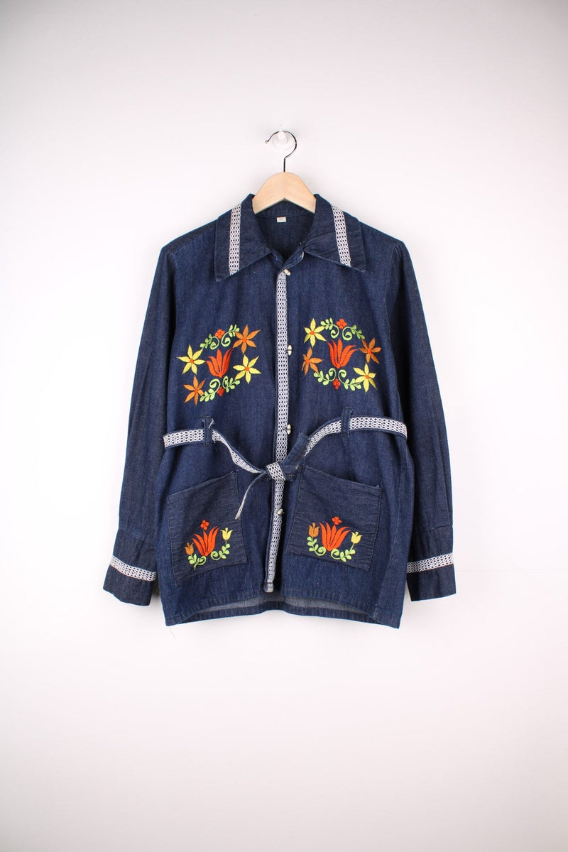 Folk Denim Jacket in a blue colourway, button up with double pockets, has a belt, dagger collar, cross stitching throughout and floral patterns embroidered on the front. 