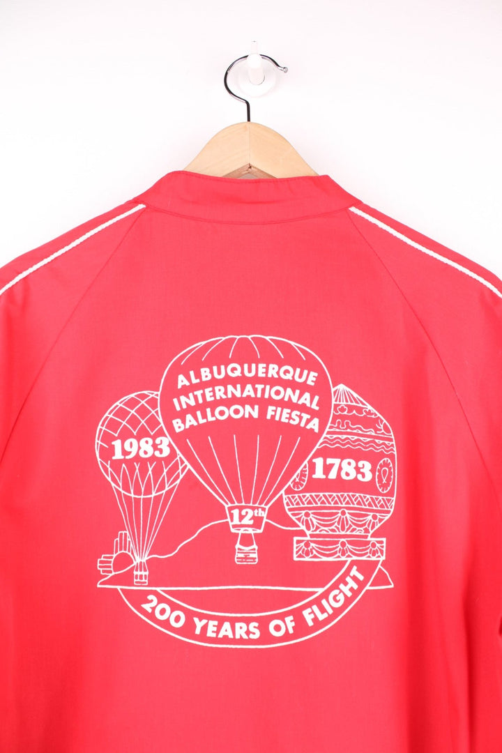 1983 Canon lightweight jacket in red with white piping, embroidered logos, a drawstring at the waist, zip closure and a back graphic of a hot air balloon and text reading 'Albuquerque International Balloon Fiesta - 200 Years of Flight'. 