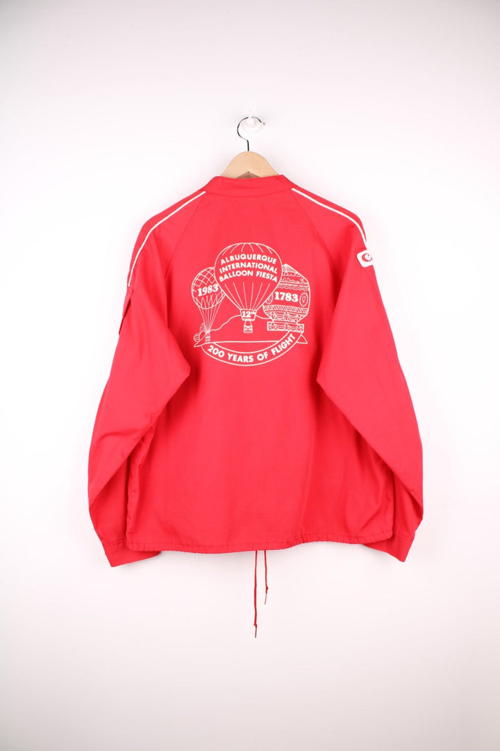 1983 Canon lightweight jacket in red with white piping, embroidered logos, a drawstring at the waist, zip closure and a back graphic of a hot air balloon and text reading 'Albuquerque International Balloon Fiesta - 200 Years of Flight'. 