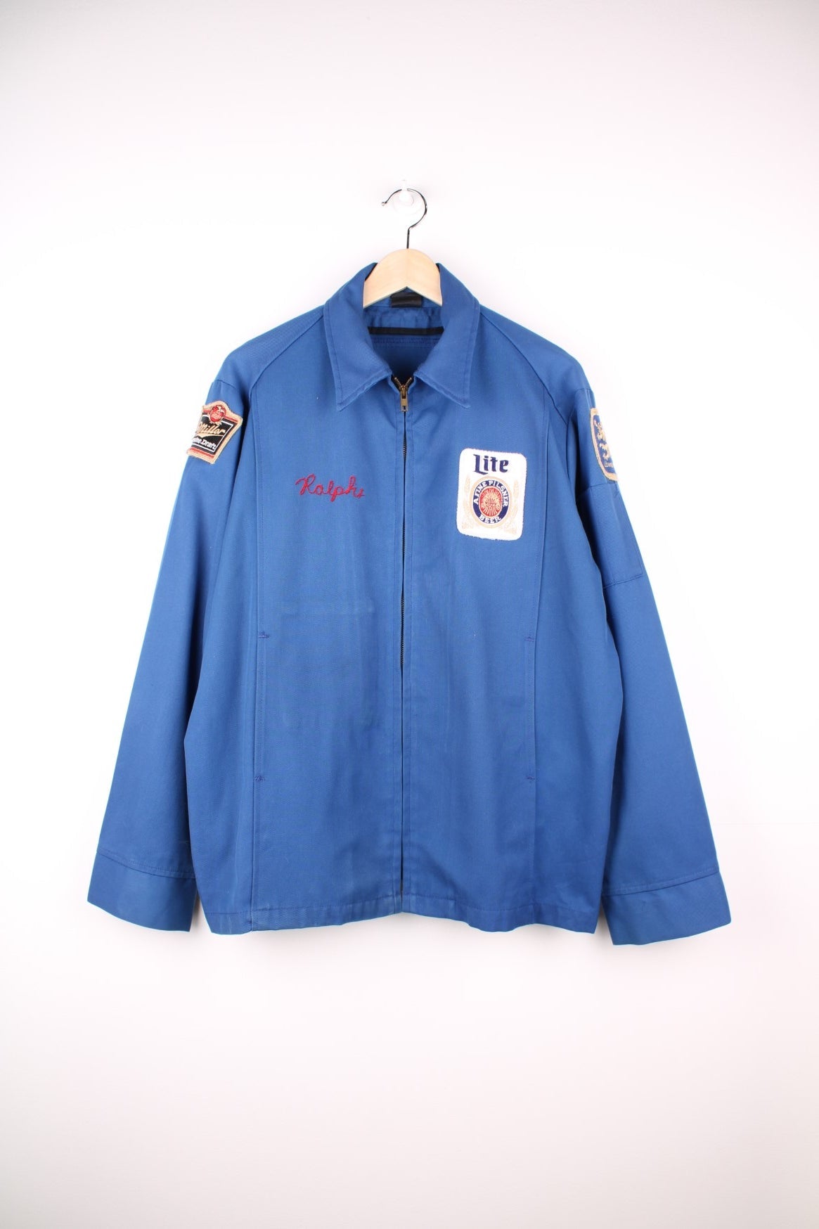 60s Miller beer lightweight delivery jacket in blue with assorted Miller beer patches on the back, ches, and sleeves, zip closure, two pockets, and the name Ralph embroidered in red on the front. Zip inside from missing detachable lining.  