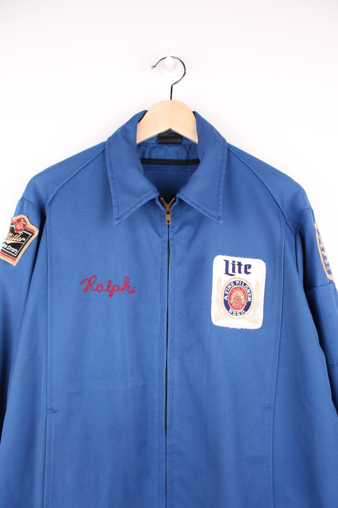 60s Miller beer lightweight delivery jacket in blue with assorted Miller beer patches on the back, ches, and sleeves, zip closure, two pockets, and the name Ralph embroidered in red on the front. 