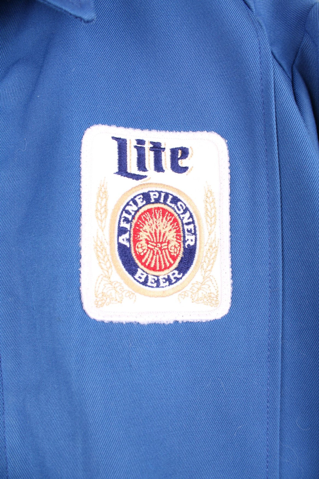 60s Miller beer lightweight delivery jacket in blue with assorted Miller beer patches on the back, ches, and sleeves, zip closure, two pockets, and the name Ralph embroidered in red on the front. 