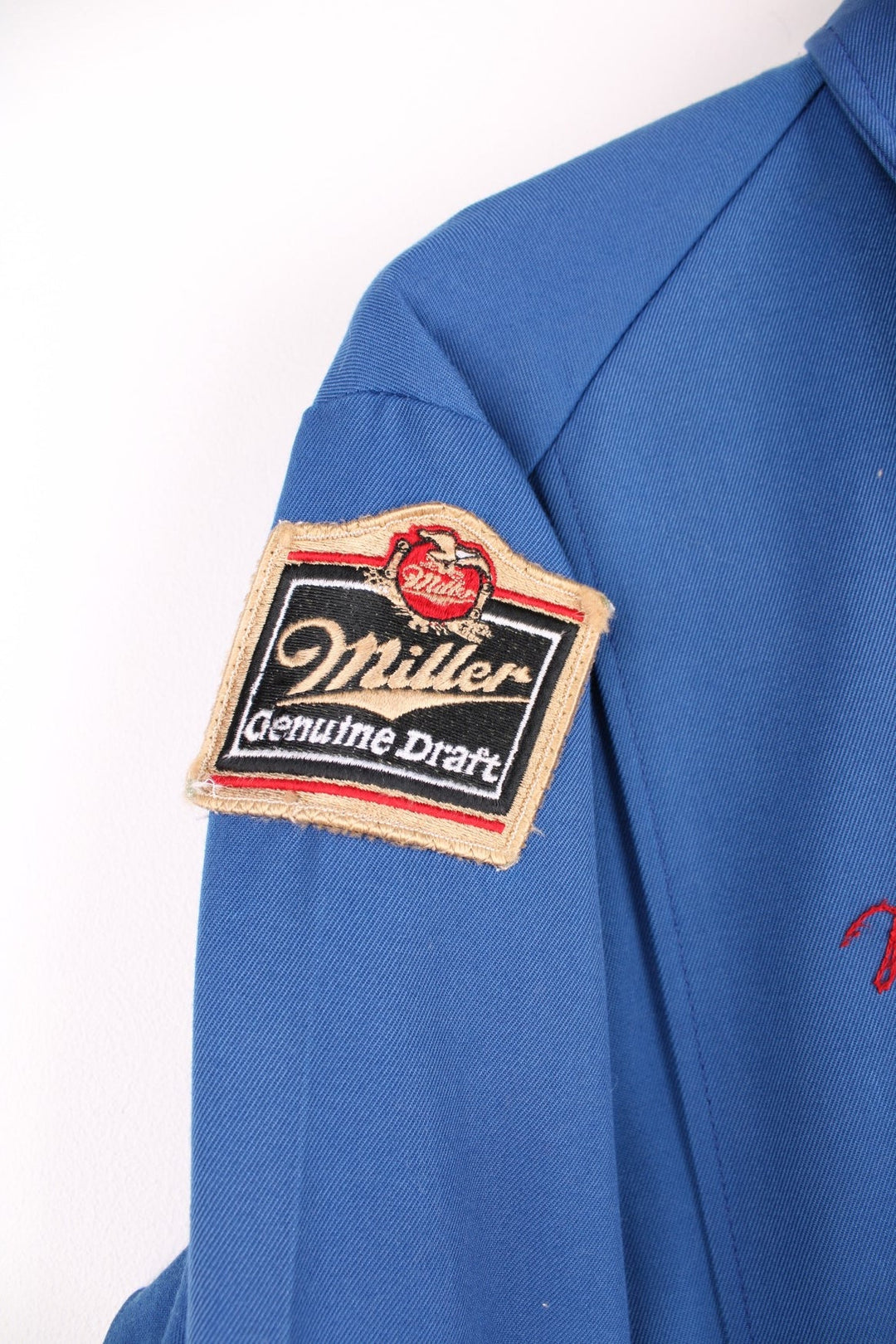 60s Miller beer lightweight delivery jacket in blue with assorted Miller beer patches on the back, ches, and sleeves, zip closure, two pockets, and the name Ralph embroidered in red on the front. 