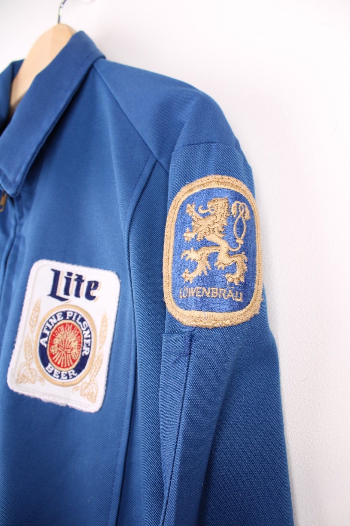 60s Miller beer lightweight delivery jacket in blue with assorted Miller beer patches on the back, ches, and sleeves, zip closure, two pockets, and the name Ralph embroidered in red on the front. 