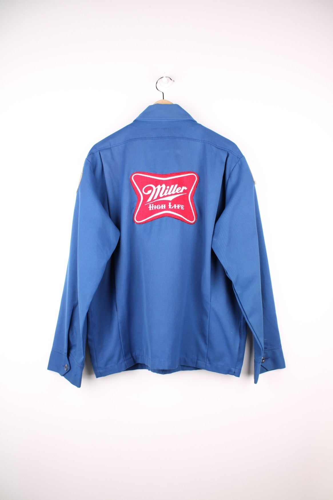 60s Miller beer lightweight delivery jacket in blue with assorted Miller beer patches on the back, ches, and sleeves, zip closure, two pockets, and the name Ralph embroidered in red on the front. 