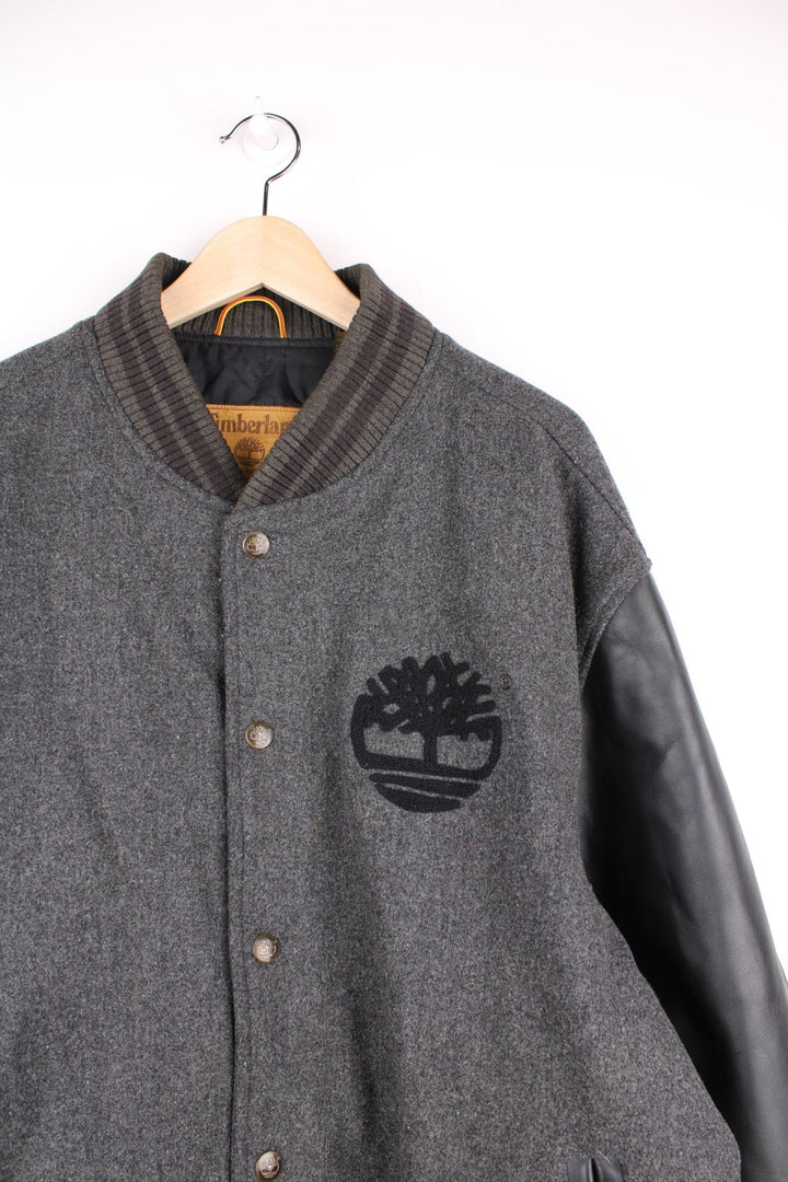 Timberland varsity jacket in a grey wool blend with 100% leather sleeves, two pockets, an embroidered tree logo on the front and spellout on the book. 