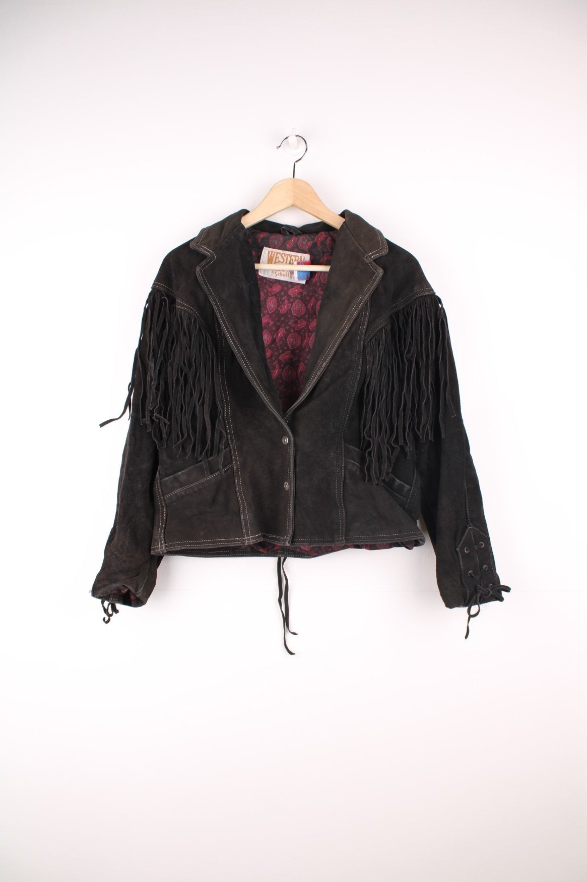 Western by Schott black suede fringe jacket with white contrast stitching, front patch pockets, button closure, a paisley lining, and lace-up detailing on the cuffs and back hem. 