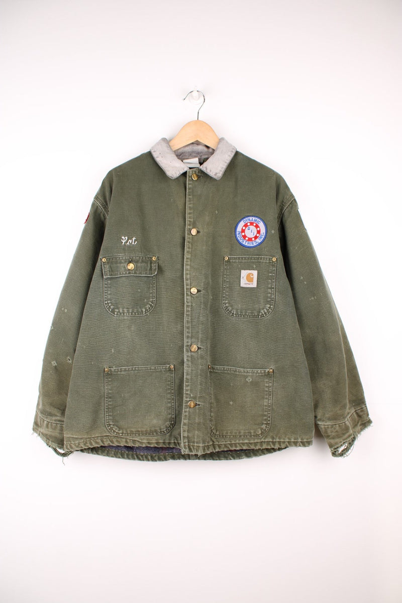 Carhartt Chore Jacket in a green colourway with a grey corduroy collar, button up with multiple pockets, blanket lining, and has the logo embroidered on the front alongside other badges. 