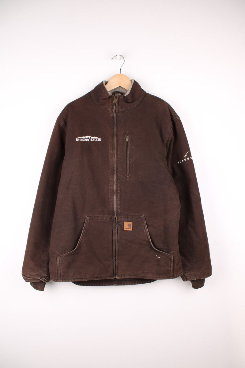 Carhartt Workwear Jacket in a brown colourway, zip up with multiple pockets, sherpa lining, and has the logo embroidered on the front alongside 'Simkins Hallin' company spell out on the front and left sleeve.  