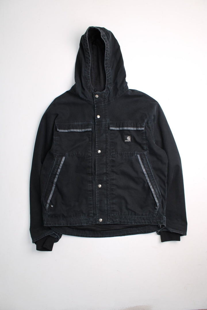 Carhartt Duck Canvas Workwear Jacket in a black colourway. Zip and button up with multiple pockets, reflective lining around the pockets, hooded, and has the logos embroidered on the front.