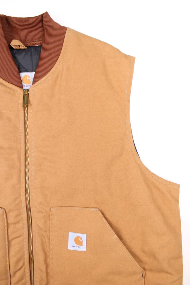 Carhartt Gilet in a tan colourway. Zips up and has side pockets, insulated with a quilted lining, and has the logo embroidered on the front.