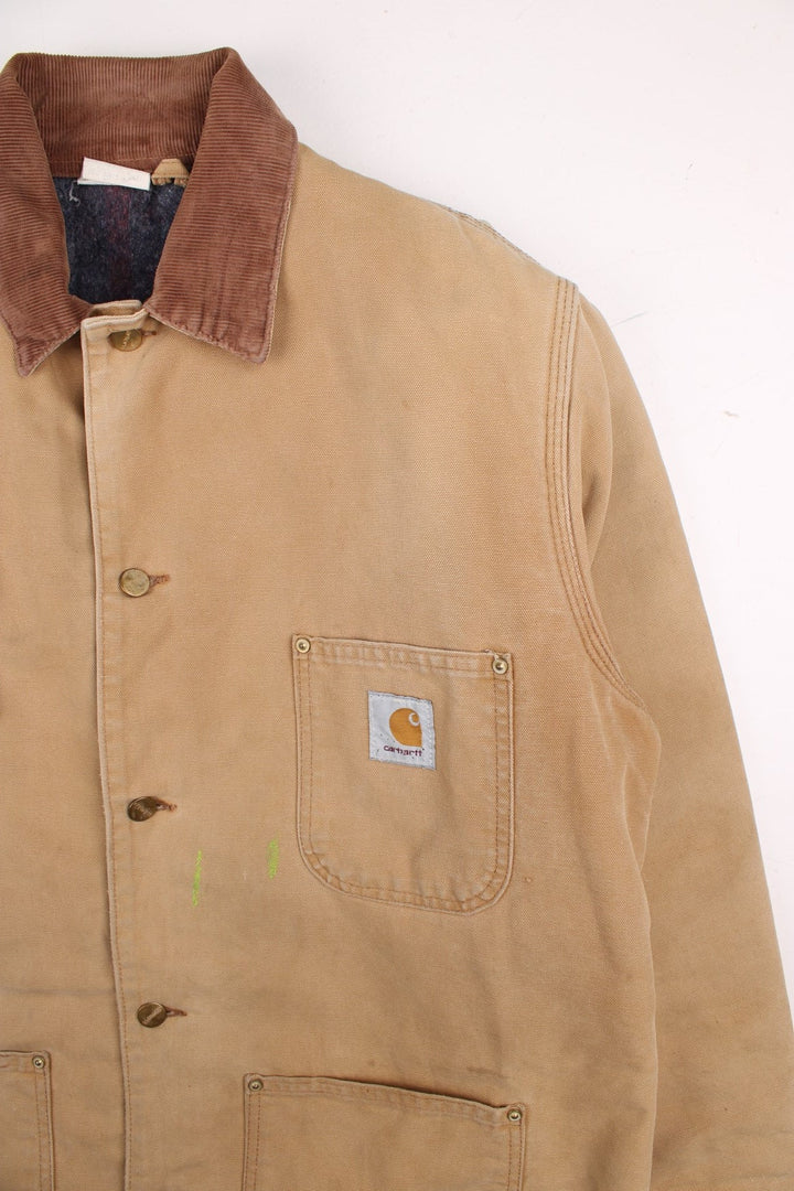 Carhartt Chore Jacket in a tan colourway with a brown corduroy collar. Buttons up and has multiple pockets, blanket lining, and has the logo embroidered on the front.