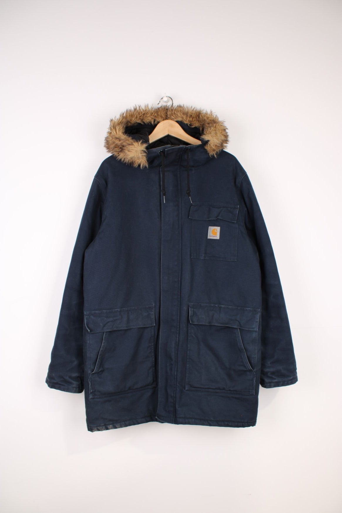 Carhartt Parka Coat in a navy blue colourway, zip up with multiple pockets, insulated with a quilted lining, has a faux fur hood, and the logo embroidered on the front. 