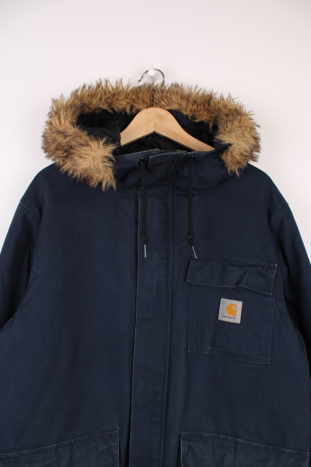 Carhartt Parka Coat in a navy blue colourway, zip up with multiple pockets, insulated with a quilted lining, has a faux fur hood, and the logo embroidered on the front. 