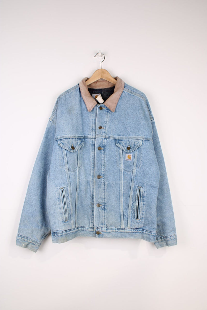 Carhartt Denim Trucker Jacket in a blue colourway with a beige corduroy collar, button up with multiple pockets, has a blanket lining, and the logo embroidered on the front. 