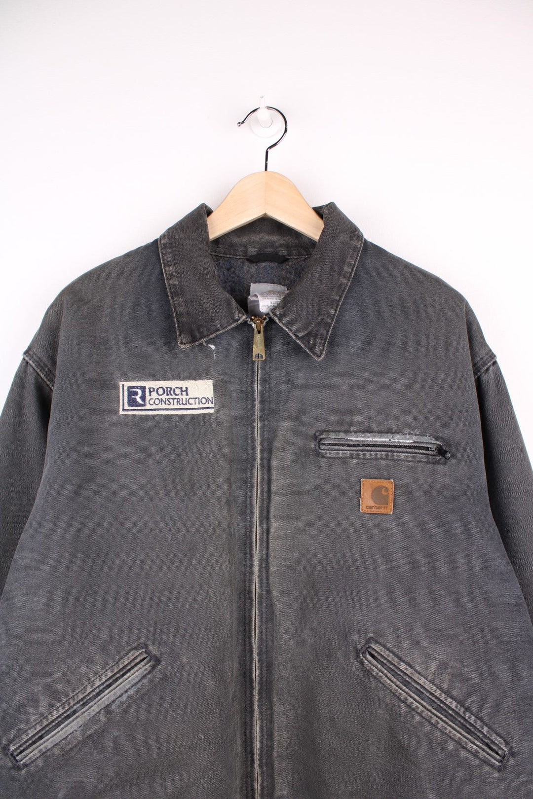 Carhart Detroit Jacket in a grey colourway, zips up and has multiple pockets, insulated with a blanket lining, and has 'Porch Construction' company spell out logo embroidered on the front alongside the Carhartt logo.