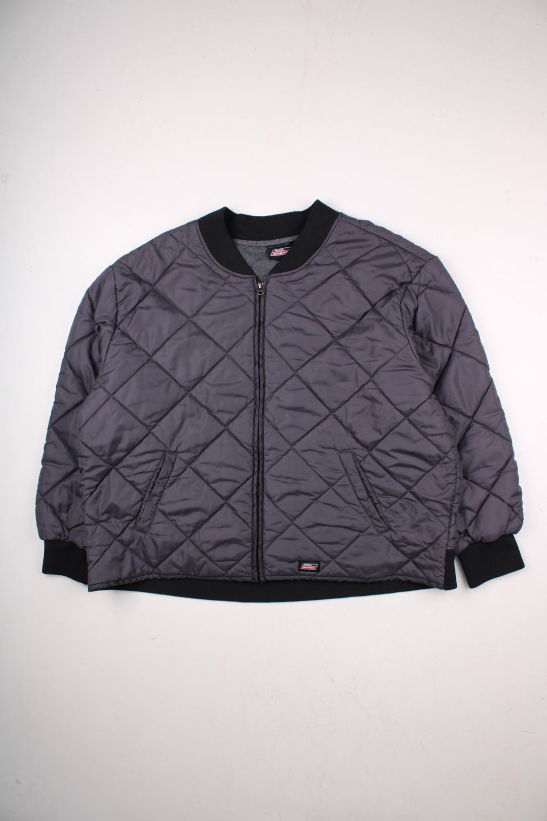 Dickies diamond quilted jacket in grey with ribbed black collar and cuffs, zip closure, two pockets, an elasticated hem, and a small logo patch on the front.