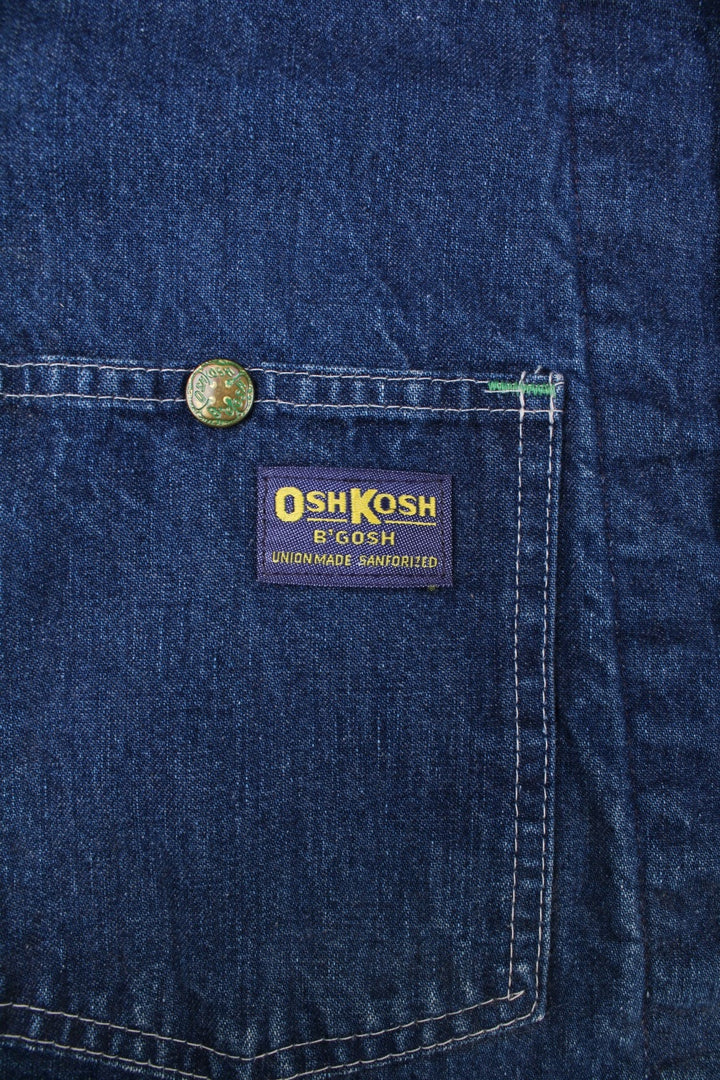 Vintage OshKosh B'Gosh sanforized denim chore jacket in blue with a red quilted lining, four patch pockets, button closure, a corduroy collar, and a small 'OshKosh B'Gosh' logo patch on the front.