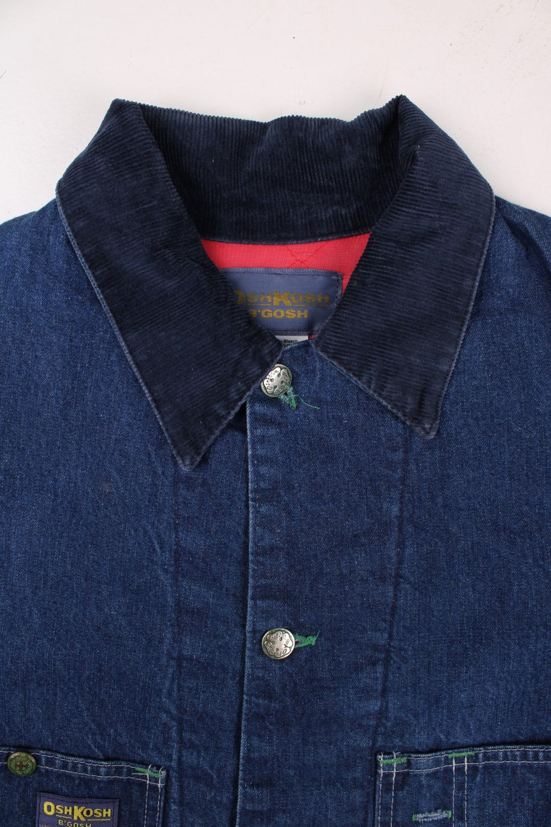 Vintage OshKosh B'Gosh sanforized denim chore jacket in blue with a red quilted lining, four patch pockets, button closure, a corduroy collar, and a small 'OshKosh B'Gosh' logo patch on the front.