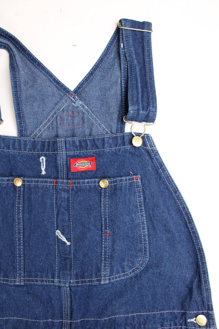 Dickies carpenter-style dungarees in blue denim with multiple pockets, white stitching, and a red logo patch. 