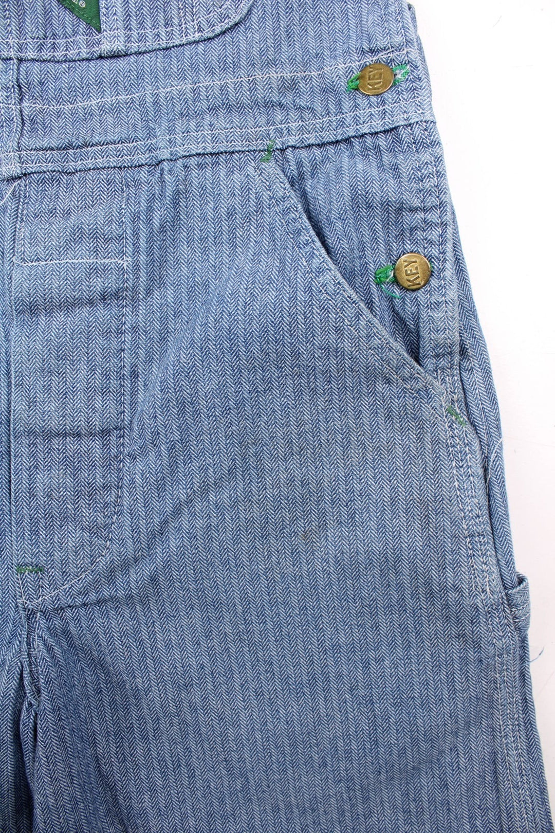 Vintage Key dungarees in blue herringbone denim with multiple pockets, branded hardware, and an embroidered logo patch on the front in green.