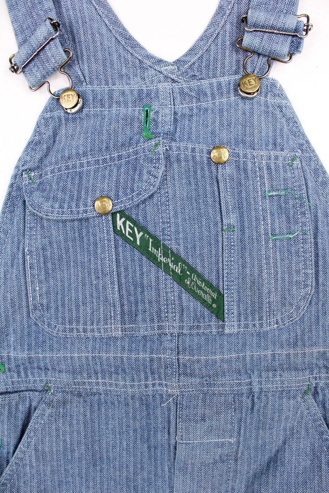 Vintage Key dungarees in blue herringbone denim with multiple pockets, branded hardware, and an embroidered logo patch on the front in green.