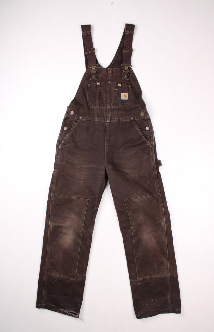 Carhartt carpenter-style dungarees in brown with multiple pockets, red-brown elasticated straps, and a white logo patch on the front.