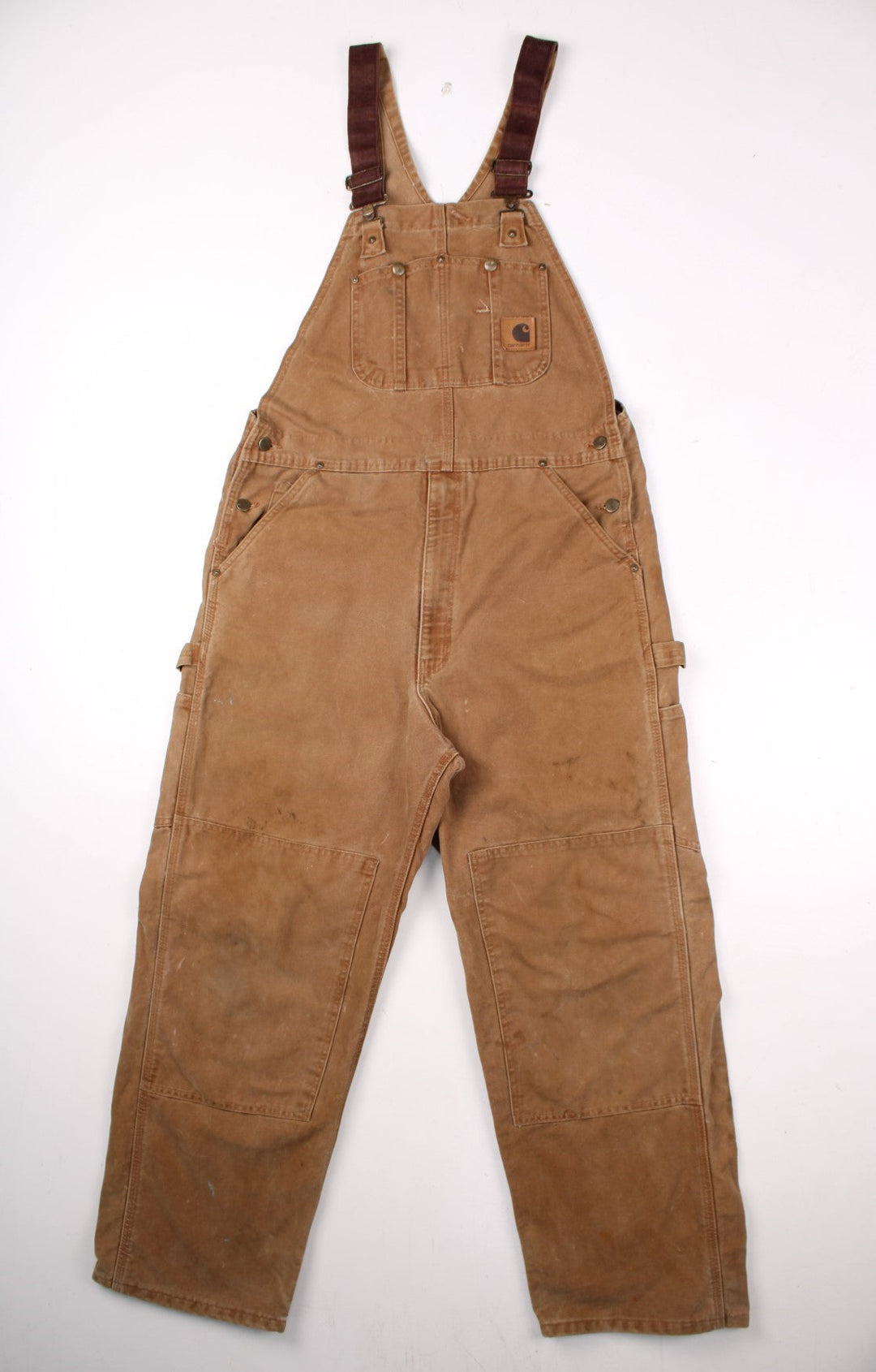 Carhartt carpenter-style dungarees in tan with multiple pockets  red-brown elasticated straps, and a leather logo patch on the front.