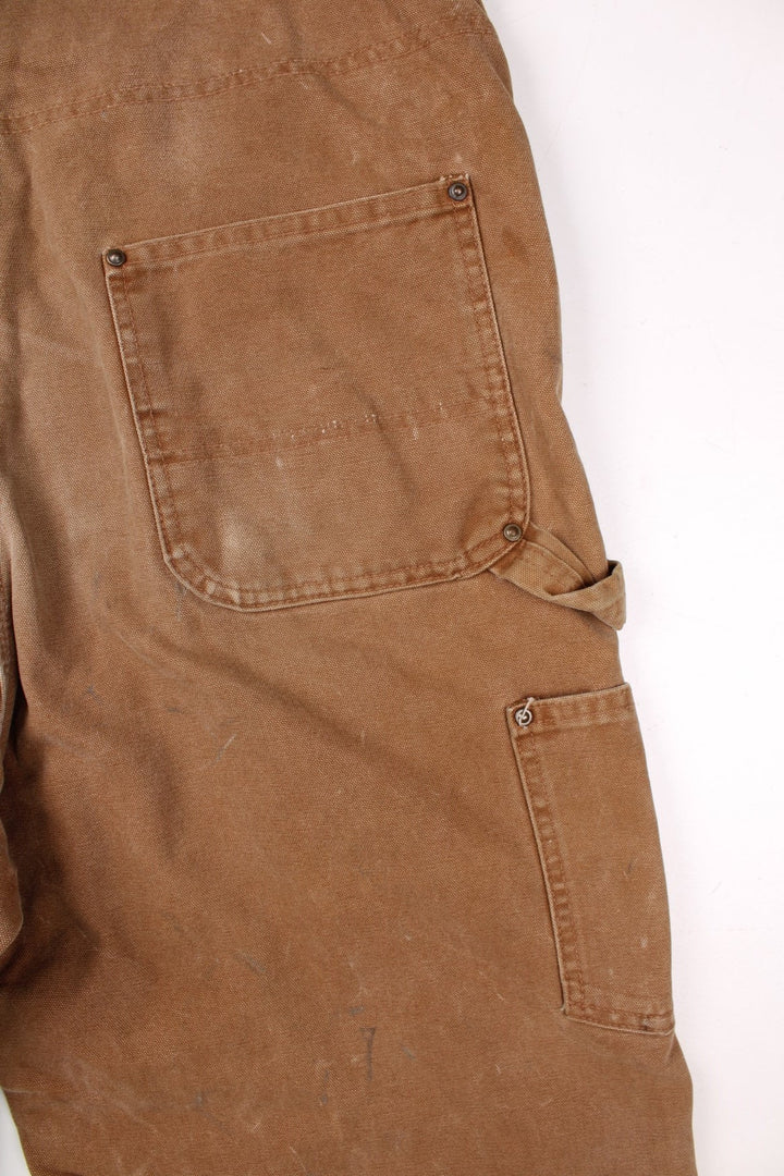 Carhartt carpenter-style dungarees in tan with multiple pockets  red-brown elasticated straps, and a leather logo patch on the front.