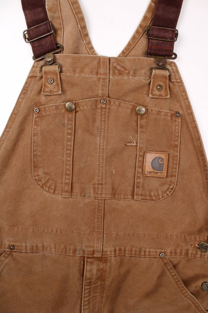 Carhartt carpenter-style dungarees in tan with multiple pockets  red-brown elasticated straps, and a leather logo patch on the front.