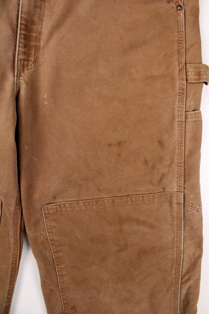 Carhartt carpenter-style dungarees in tan with multiple pockets  red-brown elasticated straps, and a leather logo patch on the front.