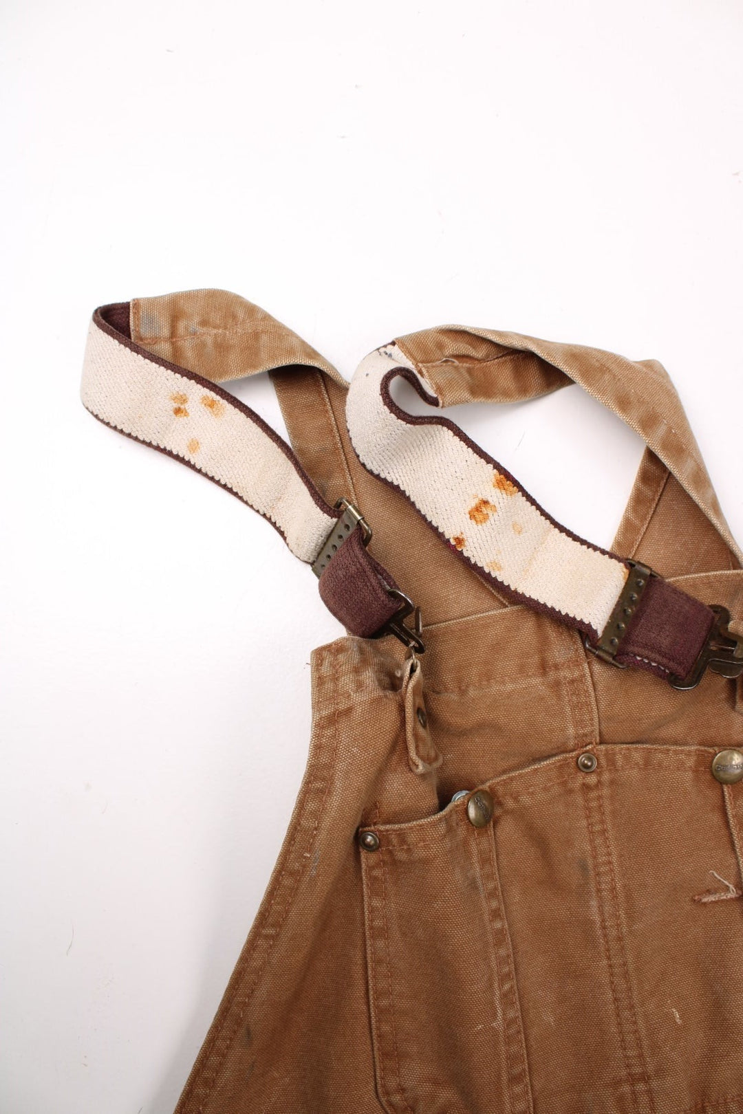 Carhartt carpenter-style dungarees in tan with multiple pockets  red-brown elasticated straps, and a leather logo patch on the front.