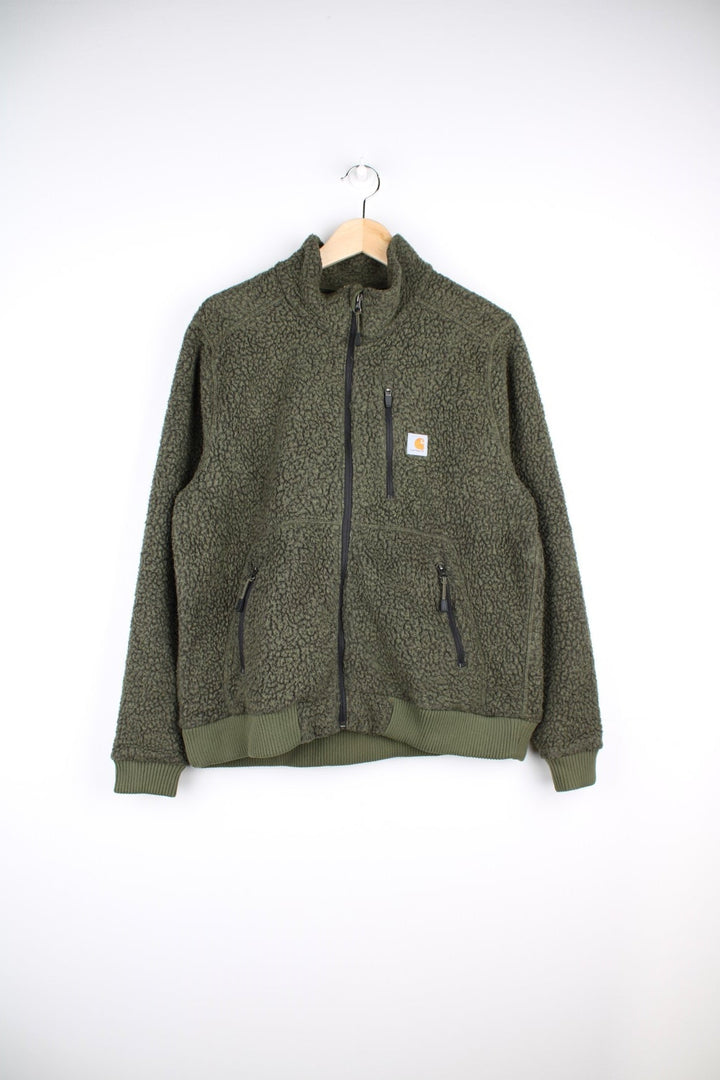 Carhartt sherpa fleece in moss green with zip closure, three zipped pockets, and a Carhartt patch on the chest.