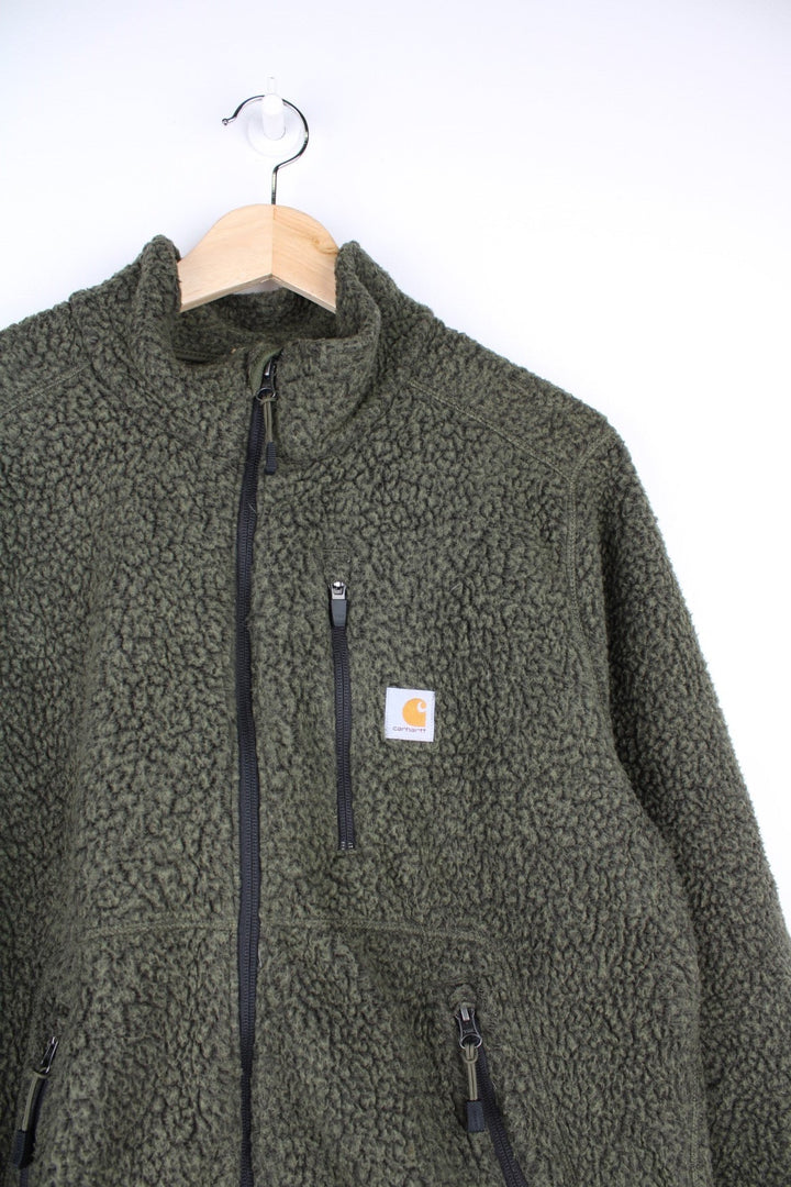 Carhartt sherpa fleece in moss green with zip closure, three zipped pockets, and a Carhartt patch on the chest.