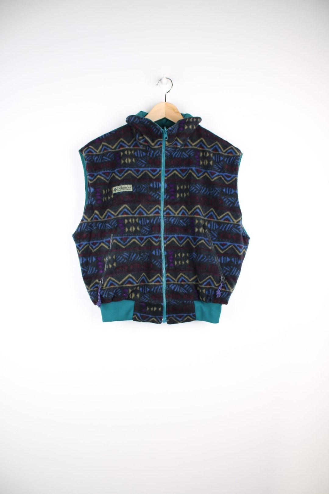 Columbia zip-through reversible fleece gilet in a blue, green, and purple geometric pattern. The reverse side is a turquoise water-resistant material. Both have pockets and a logo patch on the chest. 