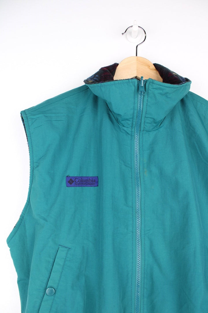 Columbia zip-through reversible fleece gilet in a blue, green, and purple geometric pattern. The reverse side is a turquoise water-resistant material. Both have pockets and a logo patch on the chest. 