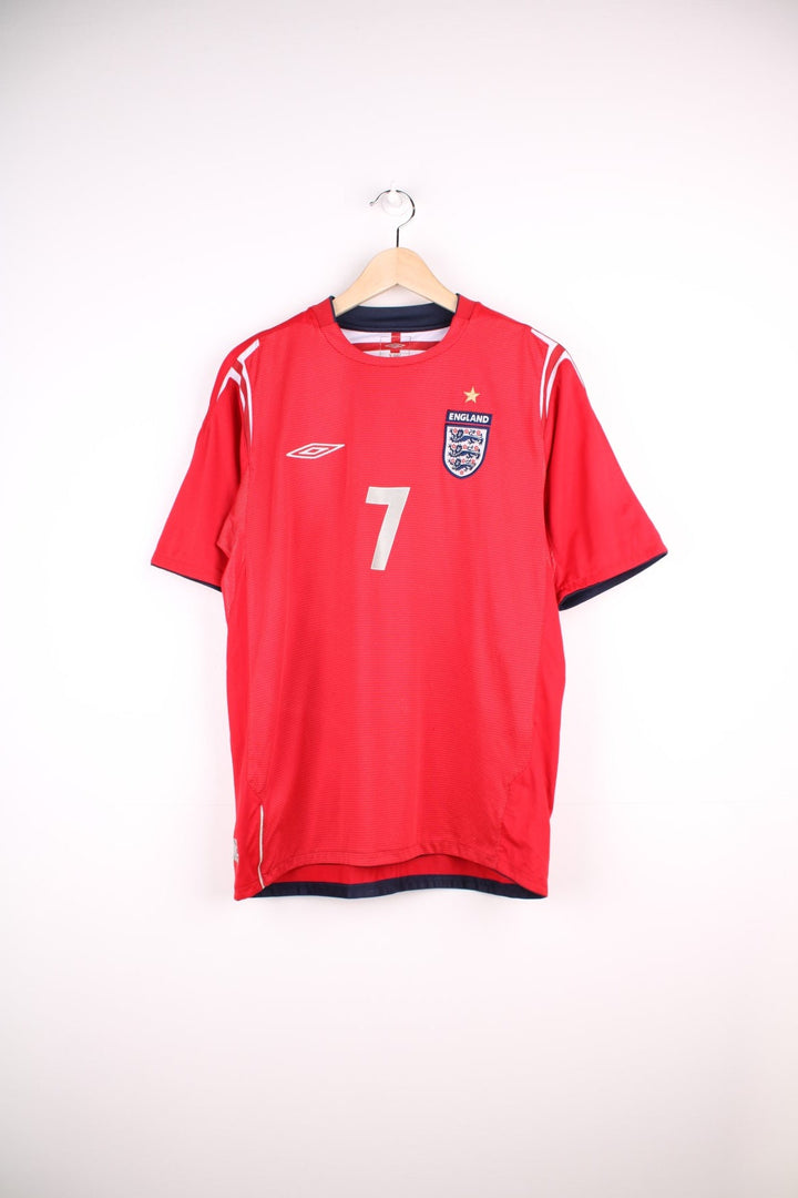 England 2004/06 Umbro Football Shirt in the red away colourway, features embroidered logo and badge on the front, and on the back has Beckham number 7 printed on.