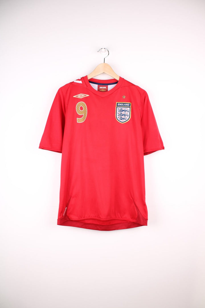 England 2006/08 Umbro Football Shirt in the red away colourway, features embroidered logo and badge on the front, and has Wayne Rooney number 9 printed on the back.