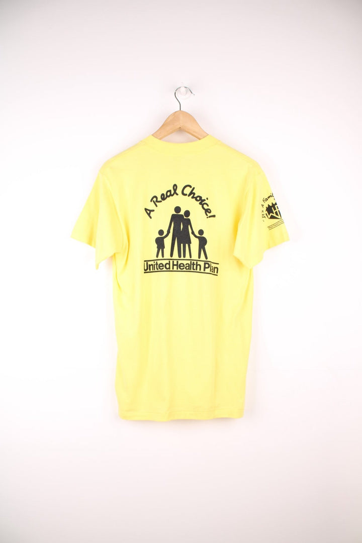 Vintage yellow graphic United Health Plan tee with cartoon design reading 'It's a Family Affair', and a Watts Health Foundation sponsorship logo on the right sleeve. 