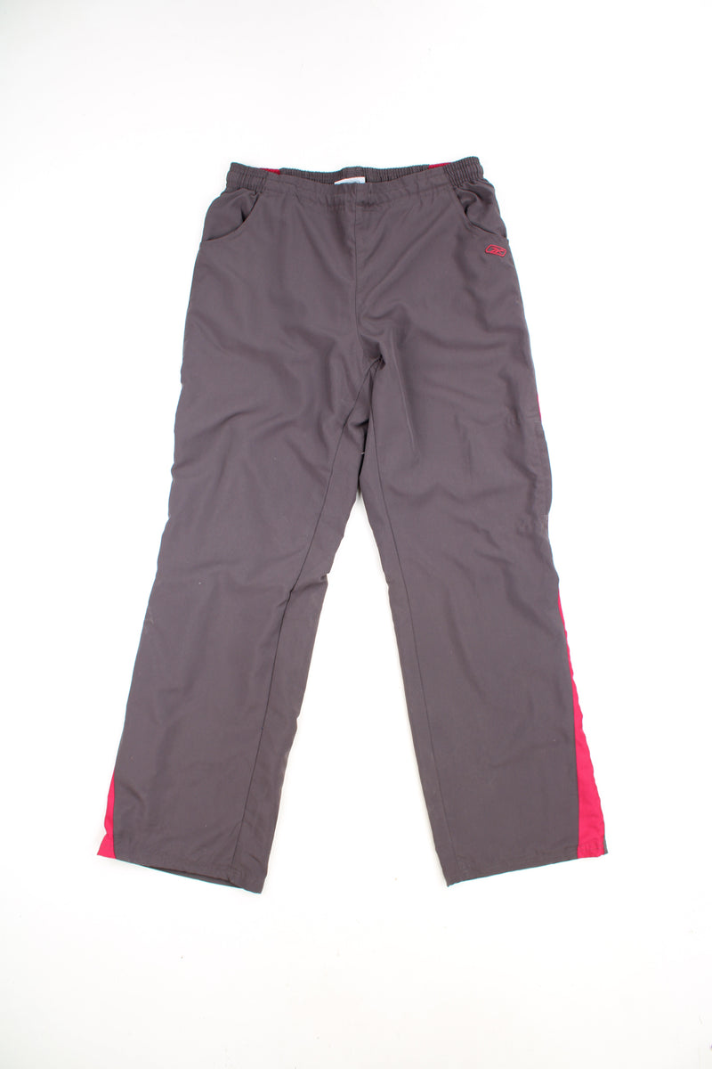 Reebok Tracksuit Bottoms in a grey and pink colourway with stripes going down the sides, adjustable waist, cargo style pocket, and has the logo embroidered on the front.