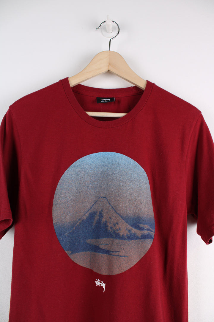 Stussy Graphic T-Shirt in a red, blue and brown colourway, has the logo printed in the centre underneath mountain graphic design.