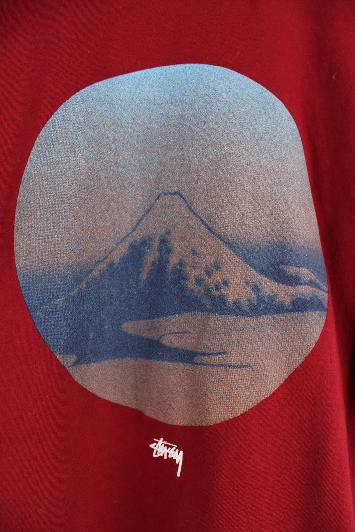 Stussy Graphic T-Shirt in a red, blue and brown colourway, has the logo printed in the centre underneath mountain graphic design.