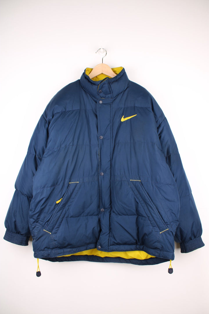 Y2K Nike Puffer Jacket in a navy blue and yellow colourway, zip and button up with side pockets, insulated and has the swoosh logo embroidered on the front and back.