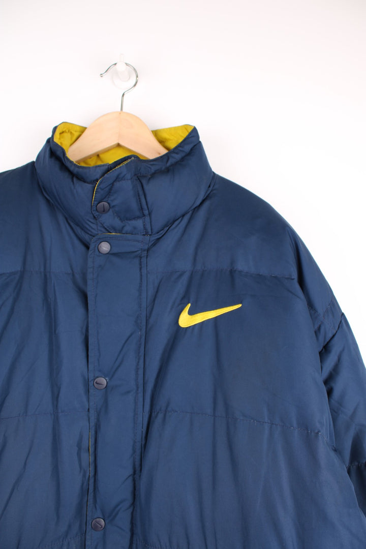 Y2K Nike Puffer Jacket in a navy blue and yellow colourway, zip and button up with side pockets, insulated and has the swoosh logo embroidered on the front and back.