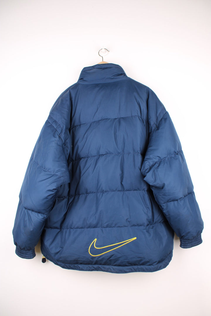 Y2K Nike Puffer Jacket in a navy blue and yellow colourway, zip and button up with side pockets, insulated and has the swoosh logo embroidered on the front and back.