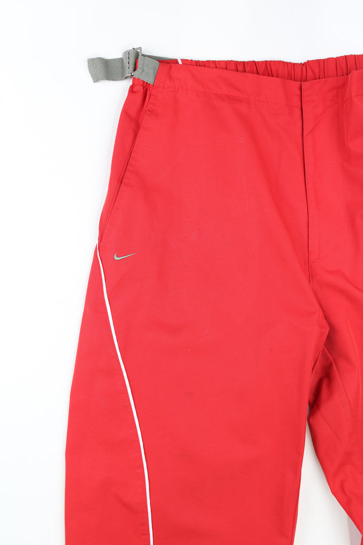 Y2K Nike red tech trouers with embroidered logo on the pocket and elasticated waistband at the back. The trousers also feature buckles at the side of the waistband to adjust the size slightly. They also have popper buttons at the cuffs to taper the leg. Size in Label: Womens L 